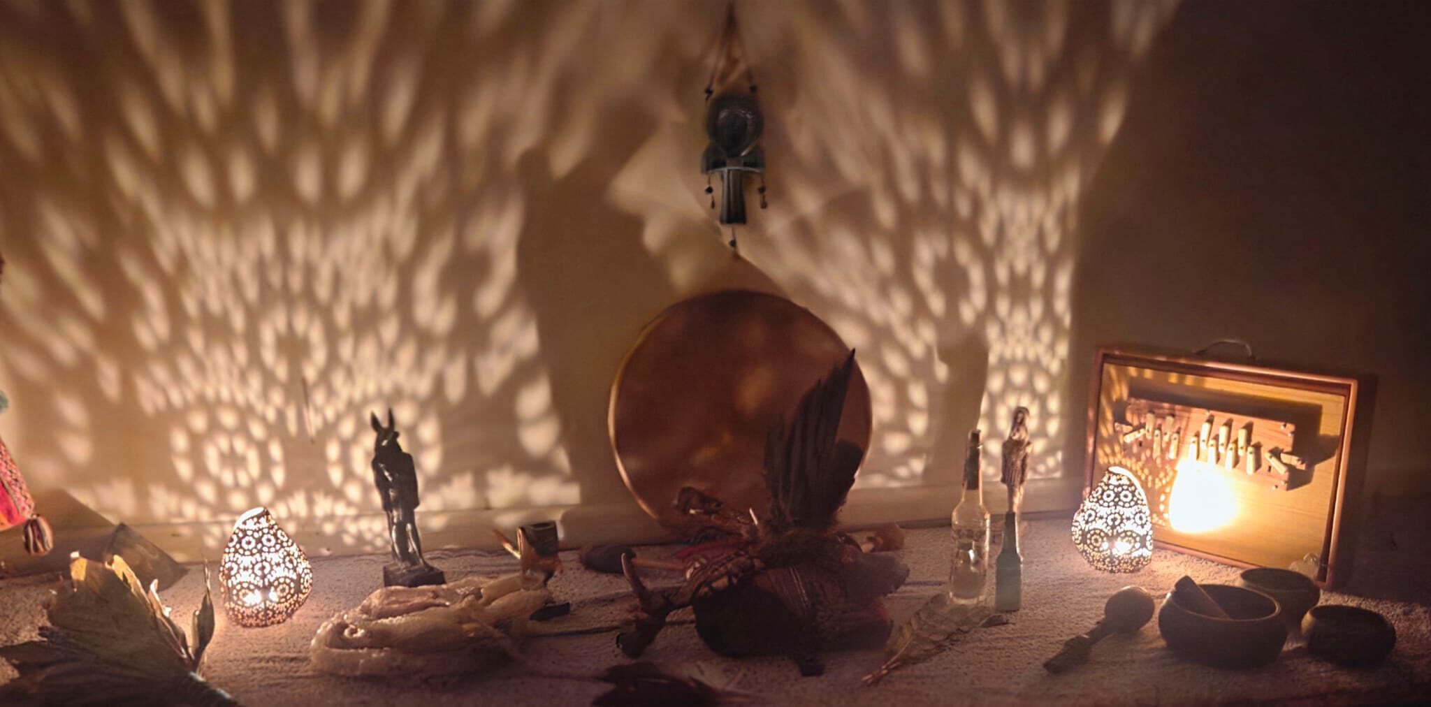 A candlelit ceremonial altar - a shamanic drum, feathers, sacred figures, and lanterns casting patterned light on the wall